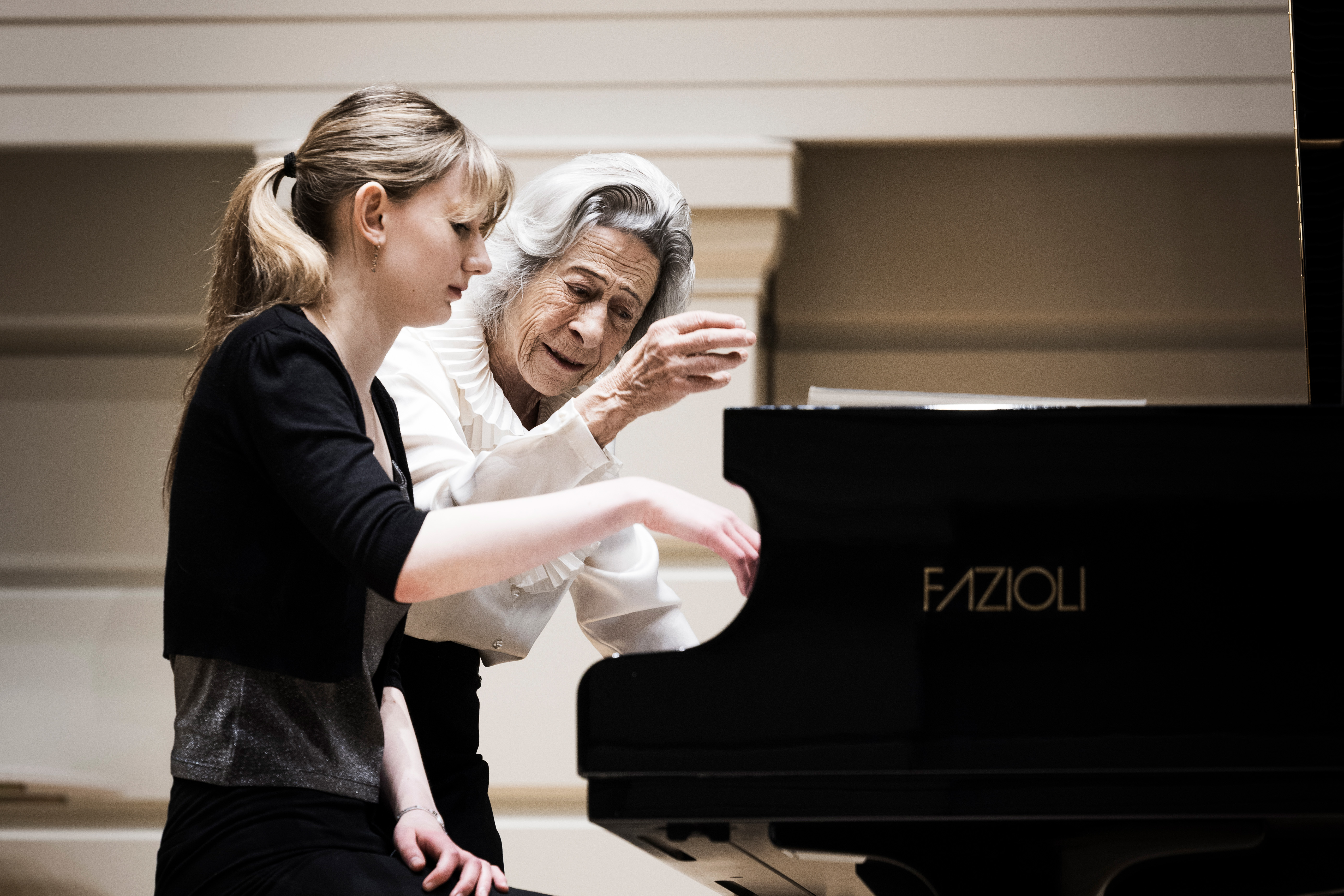 A women wearing a white shirt with grey hair, raising her hands in instruction, with a female student in smart attire, playing the piano on stage
